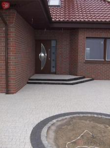 Ułożona kostka brukowa w odcieniach szarości, tworząca wzór okręgu z ziemią w środku, na tle ceglanego domu z brązowymi drzwiami i trzema schodkami wejściowymi.